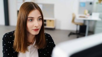 Eine junge Frau mit langen, glatten Haaren schaut konzentriert auf einen Computerbildschirm in einem modernen Büro.